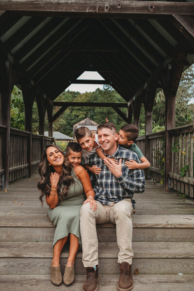 A joyful family of five laughs together on a rustic covered bridge, sharing playful moments during a lifestyle family photography session in Connecticut. Captured by Nicole Mele Photography, highlighting love, connection, and genuine happiness.