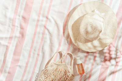 Straw sun hat, woven beach bag, and sunscreen bottle on a pink and white striped towel in bright sunlight.