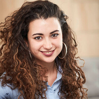 Anat has dark curly hair and large gold earrings. She is smillng.