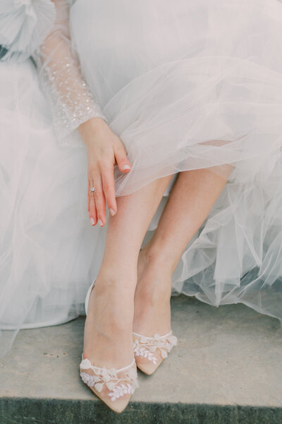 Bride seated on stone steps in a layered tulle gown, showing off designer heels — an elegant detail shot with a fashion-forward feel.