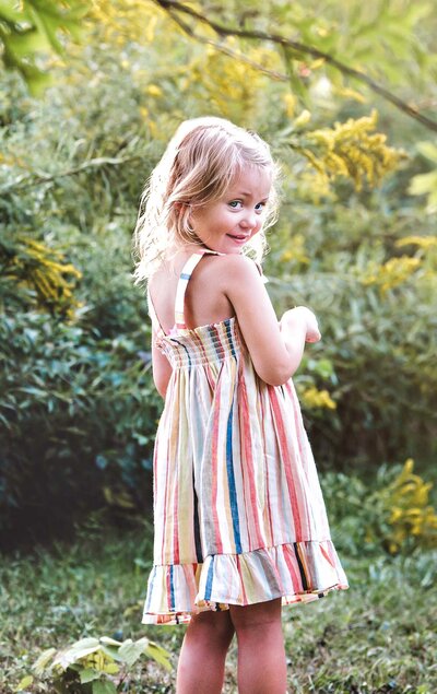 Little girl in striped dress glances over her shoulder with yellow flowers in the background..