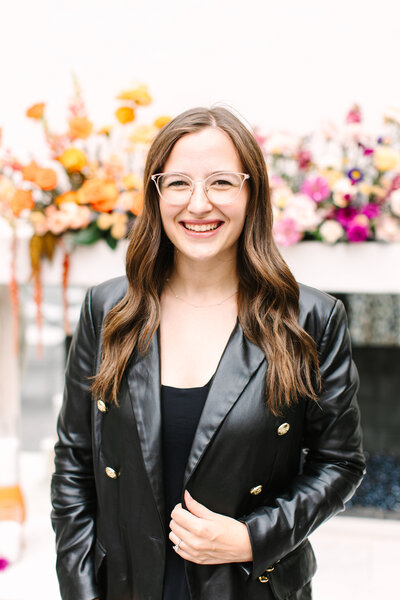 Kalie Breeze, general manager of SoHo63, a Chandler wedding and event venue, smiles while standing in front of a modern white cityscape backdrop. Her professional and friendly demeanor reflects the exceptional service and hospitality SoHo63 is known for.