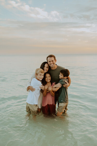 Family Photographer,  a young family of 5 huddles in shallow beach waters