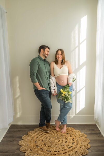 pregnant mom in white crop top and jeams holding a white and yellow boquet leaning on the wall with her husband as he gazes at her