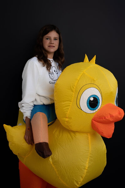 Girl wearing giant yellow inflatable duck Halloween Costume standing in front of black backdrop. 