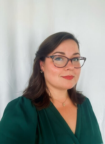 A professional portrait of Taylor White, wearing glasses and a green top, with a confident and composed expression against a neutral background.