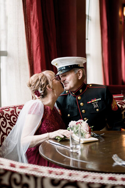 A bride in a red gown and her groom in Marine Corps Dress Blues share a quiet moment in a velvet booth.