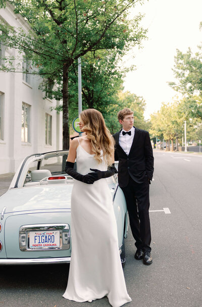 Capturing a timeless moment: bride near vintage car, groom standing behind outdoors.