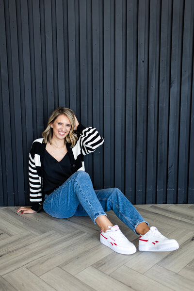 Natalia is sitting on the wooden floor in front of a black wall. She is dressed in a pair of jeans, white sneakers, a black T-shirt and a striped sweater.