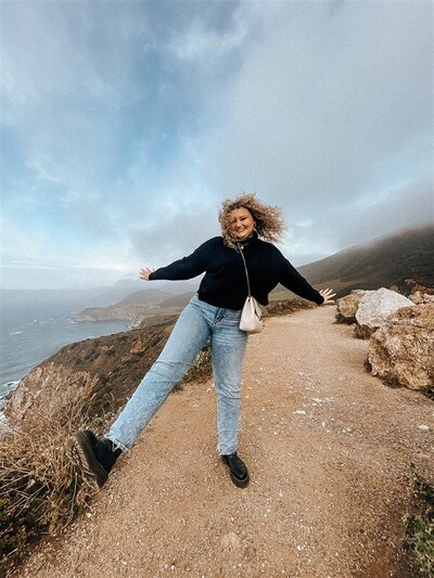 Andrea Cakmar playfully poses on a scenic cliffside trail with the ocean and misty hills in the background. She is dressed in a black sweater, blue jeans, and black shoes.