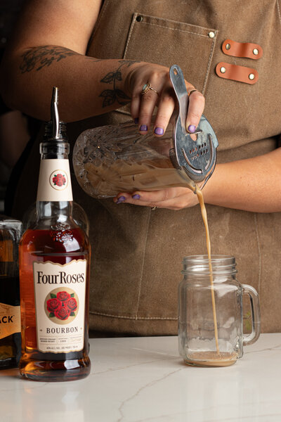 a woman pouring a coffee cocktail into a glass mason jar cup