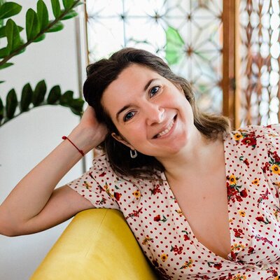 A woman with dark, shoulder-length hair, wearing a light-colored floral dress, leans on a yellow couch with a relaxed smile. The background includes green plants and decorative glass panels, creating a cozy and vibrant atmosphere.