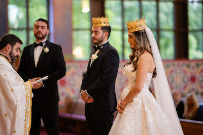 A couple wearing crowns exchanges vows during an Orthodox wedding ceremony at Saints Peter & Paul Antiochian Orthodox Christian Church in Maryland, captured by Nathan Sottung Photography as a second shooter for Justy Photography