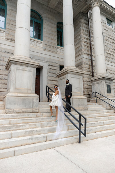Bride standing gracefully on courthouse steps, wearing a short white off-the-shoulder gown with a veil, gazing to the side.
