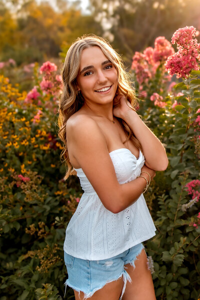 A beautiful high schoolto the side with her arm cossed and her left hand resting behind her neck, posed next to some pink flowers at sunset. senior girl standing