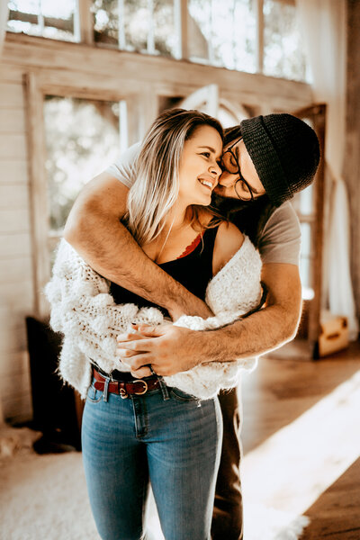 Young man with beanie on wraps up his wife from behind as he kisses her on the cheek. His beautiful wife looks back at him and smiles big knowing she is loved by him.