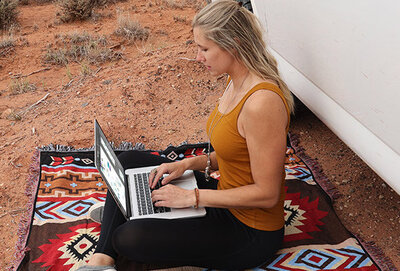 Blonde haired woman sitting outside against a van in the desert. She has a laptop on lap, working. Beside her sits a black camera, brown hat and sunglasses.