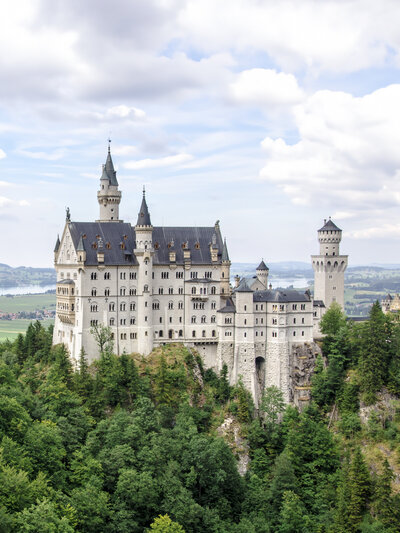 A fairytale-like castle with tall spires and a mountainous backdrop, surrounded by lush green trees. Fluffy clouds fill the sky, adding to the majestic scenery.