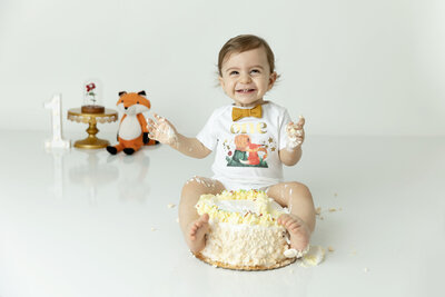 A toddler boy in a gold bowtie and white onesie wraps his feet around a yellow cake in a studio while giggling