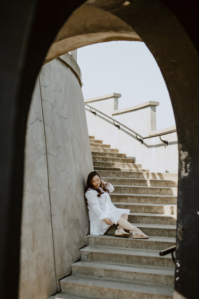 Anne, an Asian woman, is sitting in the curved corner of the Stanley Park Lighthouse with her legs slightly bent and looking down while candidly adjusting her hair with her left hand and the ruffle of her white knee length dress with her right hand. She is wearing suede espadrilles.