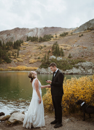 bride and groom are hiking up the trail with trees surrounding them. the bride is looking back at the groom as he is holding onto his backpack. the bride has flowers in her hair and is holding her dress up to reveal that she is wearing hiking boots.