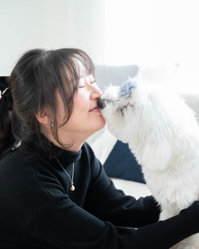 A woman with dark hair in a ponytail, wearing a black turtleneck, smiles with her eyes closed as a fluffy white dog gently licks her nose. The dog has a soft blue flower accessory on its head. They are sitting on a light-colored couch in a bright, sunlit room, sharing a joyful and tender moment during an in-home session by Calgary pet photographer Chase Magic Photography.