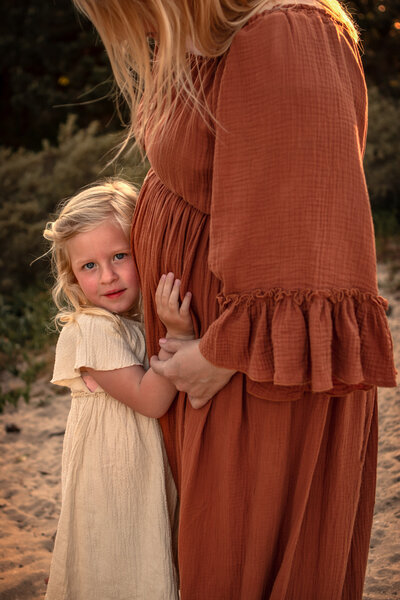 Meisje knuffelt zwangere buik moeder tijdens een zwagerschaps fotoshoot in de duinen van Noordwijk