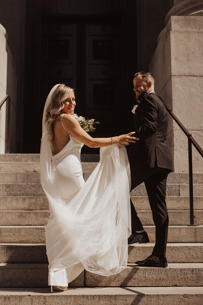 Bride and groom smiling on stone steps outside a building, bride with a trailing veil.