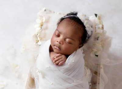 Rochel Konik Photography | Top NYC Brooklyn Newborn Photographer captures baby girl wrapped in white tulle with pearl embellishments. Baby's hands are peeking out of the wrap and resting under baby's chin. Baby is sleeping and wearing a white headband.