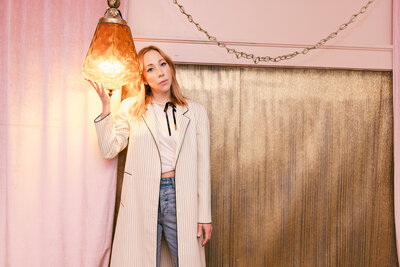 Editorial-style branding portrait of a woman in a long striped coat holding an amber pendant light, standing against a velvet pink and gold backdrop — photographed in Greenville, SC with a creative, vintage aesthetic.