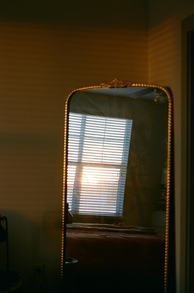 A vintage mirror reflects a window with blinds, with warm sunlight streaming through, casting serene shadows in a cozy room.