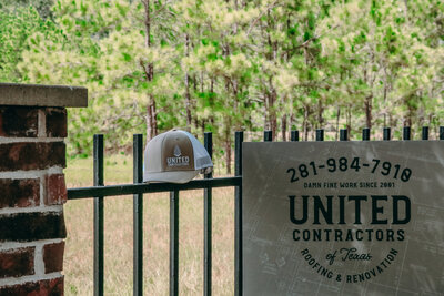 A red brick column and beautiful wrought iron fence sits before a forest full of trees. A United Contractors of Texas hat sits on the fence next to a sign featuring their logo and phone number.