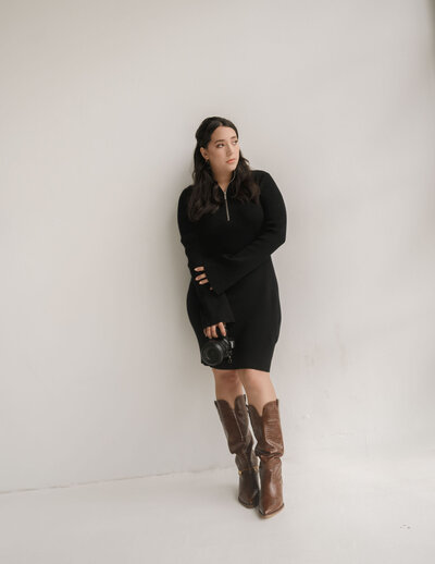A woman in a black dress, holding a camera, leans against a white wall. She wears knee-high brown boots, exuding a contemplative and relaxed vibe.