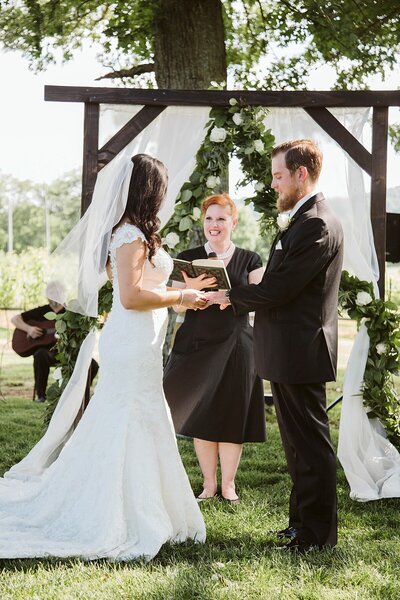 Officiant in black marrying a bride in a lace trumpet gown and groom in a black suit under and arbor with white flowers at Arrington Vineyard