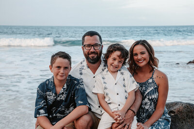 family of four sitting on a rock at ironwoods beach in Maui with the ocean in their backdrop all wearing white and blue colors