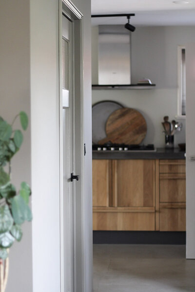Moderne keuken met houten kastdeuren, donkere details en een strakke opstelling, gezien door een lichte hal met groene accenten.