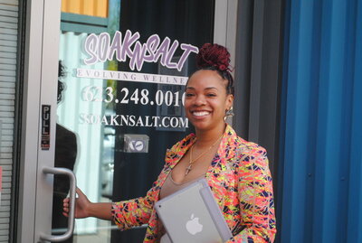 A smiling woman with braided hair and a colorful blazer holds a MacBook and stands at the entrance of SOAKNSALT, a wellness business in Tolleson, Arizona, about to open the glass door for her next message therapy session.