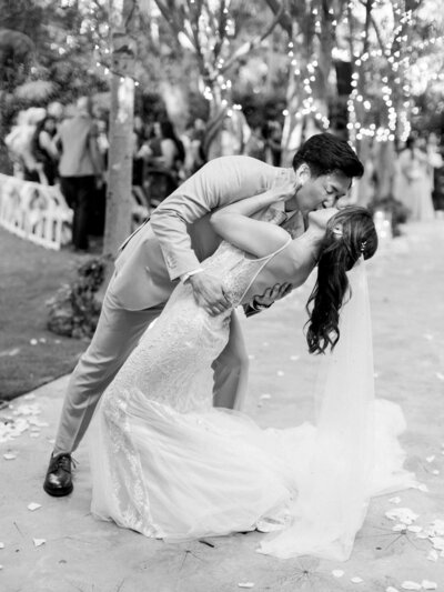 A couple in elegant attire shares a romantic dance dip and kiss at an outdoor wedding, surrounded by guests and twinkling lights.