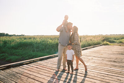 Mom and Dad kissing with baby on his shoulders and little boy standing in front of them