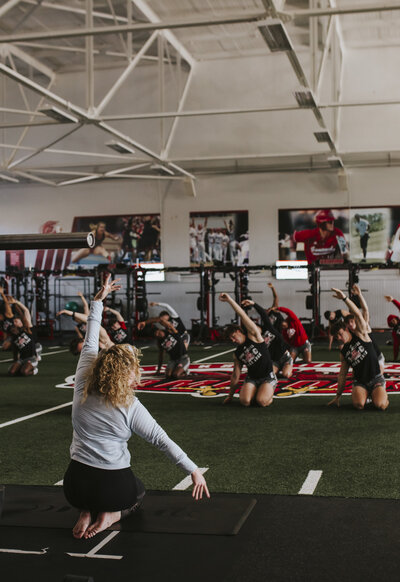 Color image of Alex Paige Yoga teaching a yoga class to a group of male football players in a gym with a turf