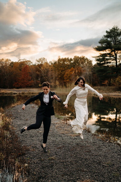 Wedding couple jumping in front of lake and trees at sunset in Hudson Valley