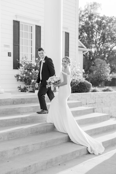 Black and white photo of a bride and groom walking up stone steps in front of a white Southern-style venue with columns, as the bride glances back with a smile. Taken in Denton, Texas.