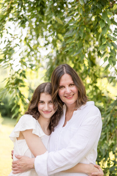 Mom and daughter hug each other while looking and smiling at the camera.