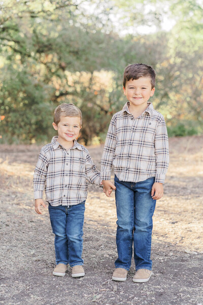Two little boys stand on a sandy path holding hands for their family portraits in Austin, TX. They wear matching plaid shirts and blue jeans.