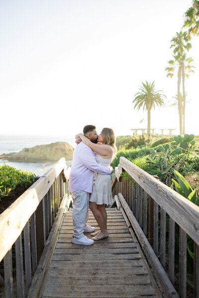 engaged couple hug on a bridge above the beach