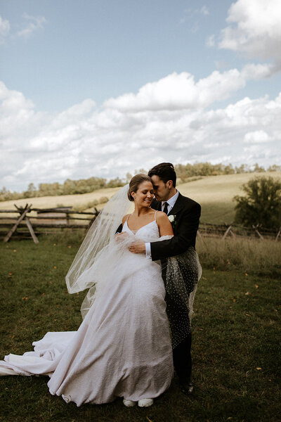 A couple in wedding attire embraces outdoors in a grassy field with a wooden fence and cloudy sky. Captured through the artful lens of Toronto destination wedding photographer, Jennifer van Son at Farmill Weddings in Peterborough, the bride wears a veil while the groom stands nearby in his suit, exuding timeless elegance.