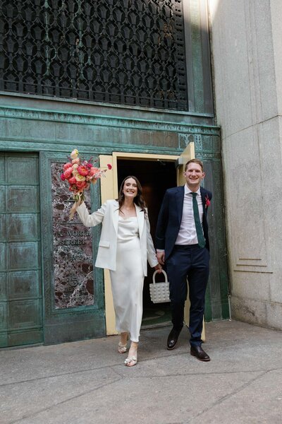 nyc city hall elopement couple leaving city hall exit with beaded bag and bloom bloom bouquet