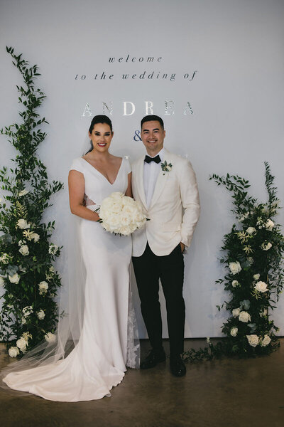 A bride in a white dress and a groom in a white tuxedo jacket pose together, smiling, in front of a stunning wedding backdrop with green foliage and white flowers—a testament to the excellence provided by their full service wedding planning team.