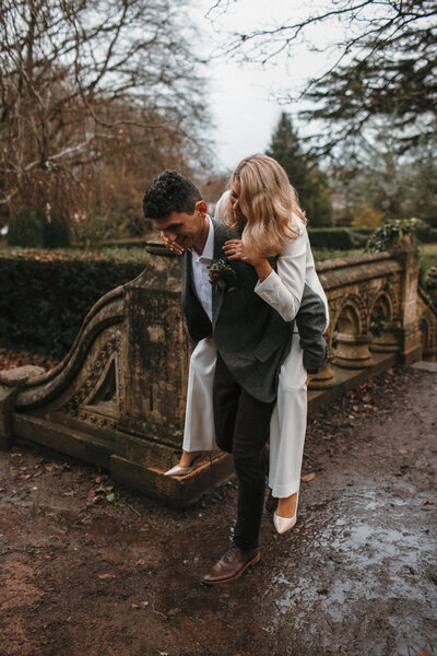Bride in white suit is carried by her groom over muddy puddles at winter civil ceremony