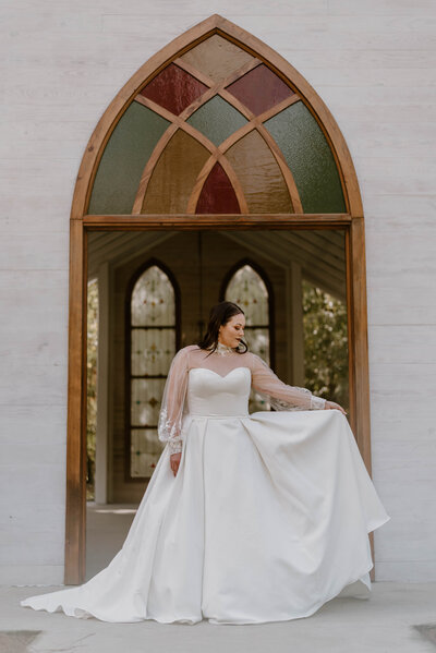 A beautiful portrait of a bride fanning out her dress standing in front of a stained glass door at Cypress Grove Open Air Chapel in Louisiana. 